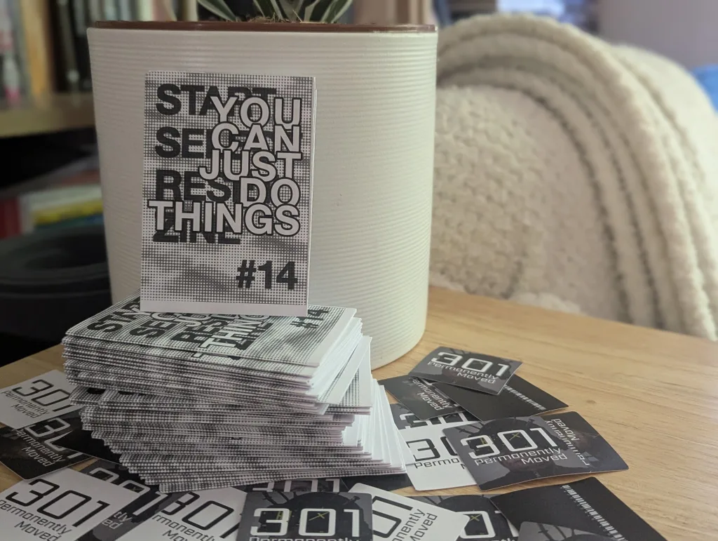 You Can Just Do Things #14 zines and 301 Permanently Moved stickers on a wooden table next to a white ribbed plant pot.