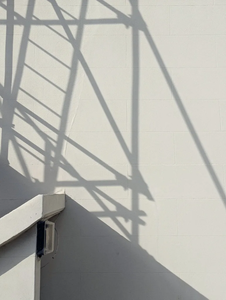 Scaffolding shadows create a geometric pattern on a white tiled wall beside a concrete staircase and a black outdoor light.