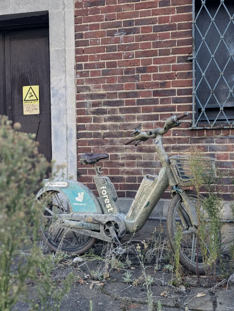 A mud-caked Forest e-bike with Deliveroo branding parked against a red brick wall next to a dark door with a yellow "Danger of Death" sign.