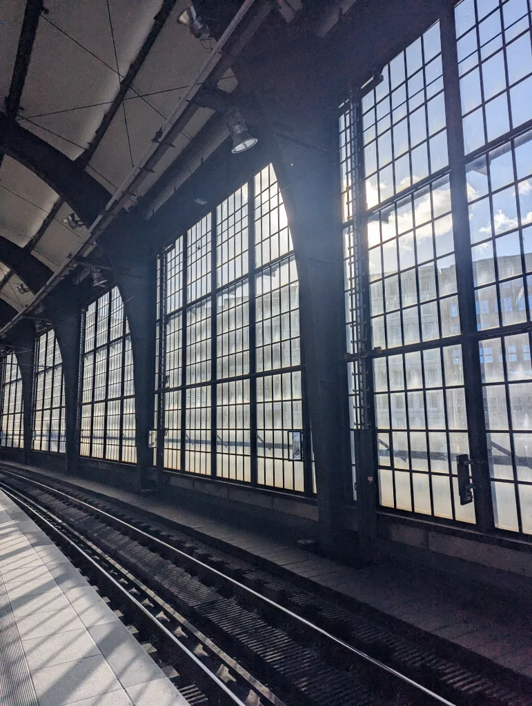 Sunlight streaming through large industrial grid windows onto empty train tracks and a tiled platform inside a station.