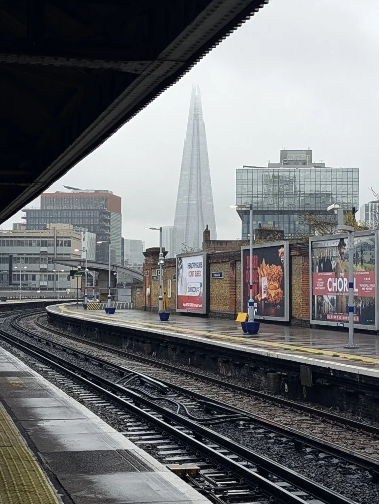 The Shard looms through the mist beyond a wet London train platform lined with billboards under a grey sky.