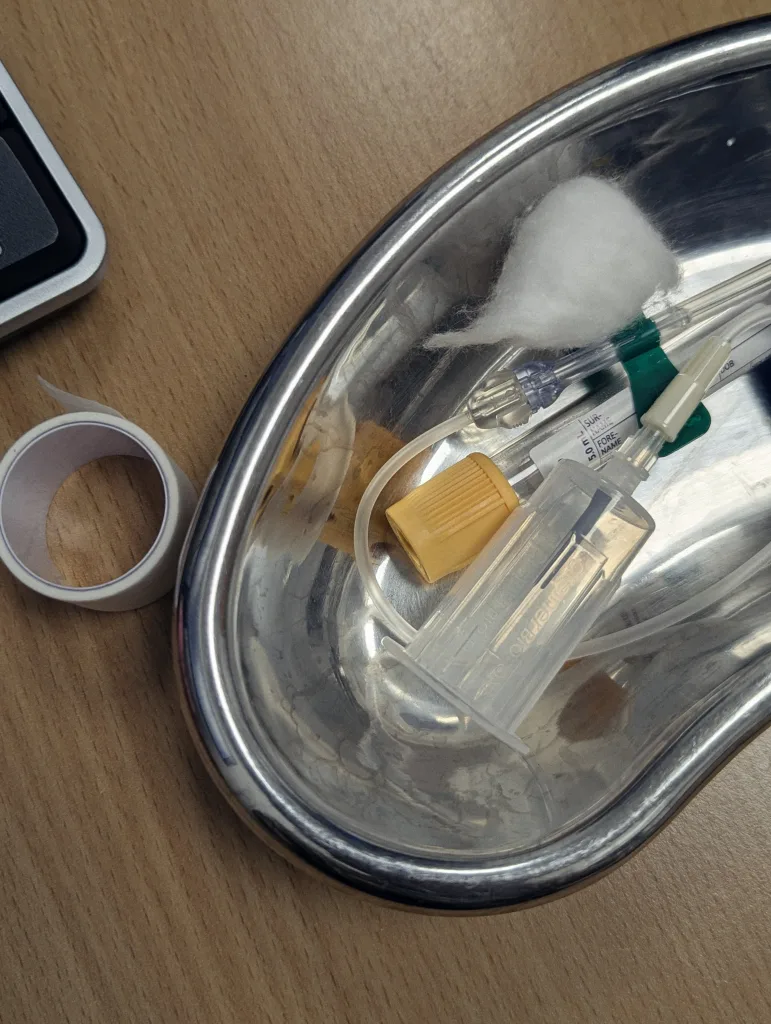 Medical supplies including an IV line and cotton ball in a metal kidney dish on a wooden desk, next to a roll of white medical tape.