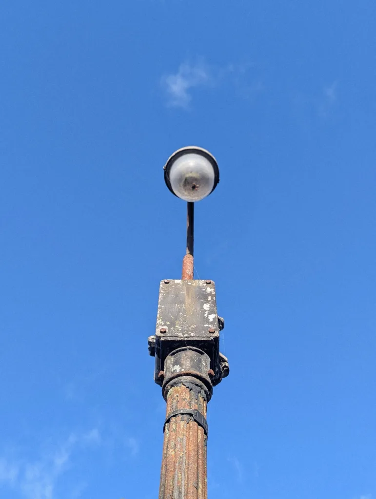 Upward view of a vintage, rusted iron lamppost and its electrical box against a bright blue sky.