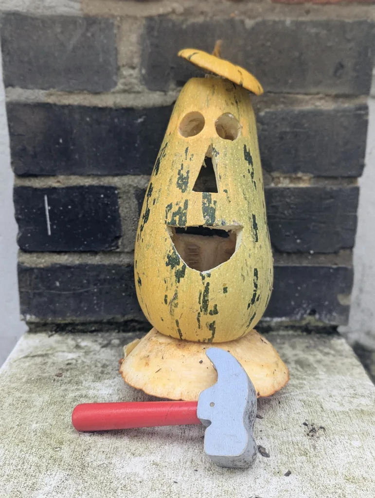 A tall yellow-green squash carved with a smiling face and lid hat sits beside a toy hammer on a stone ledge against a dark brick wall.