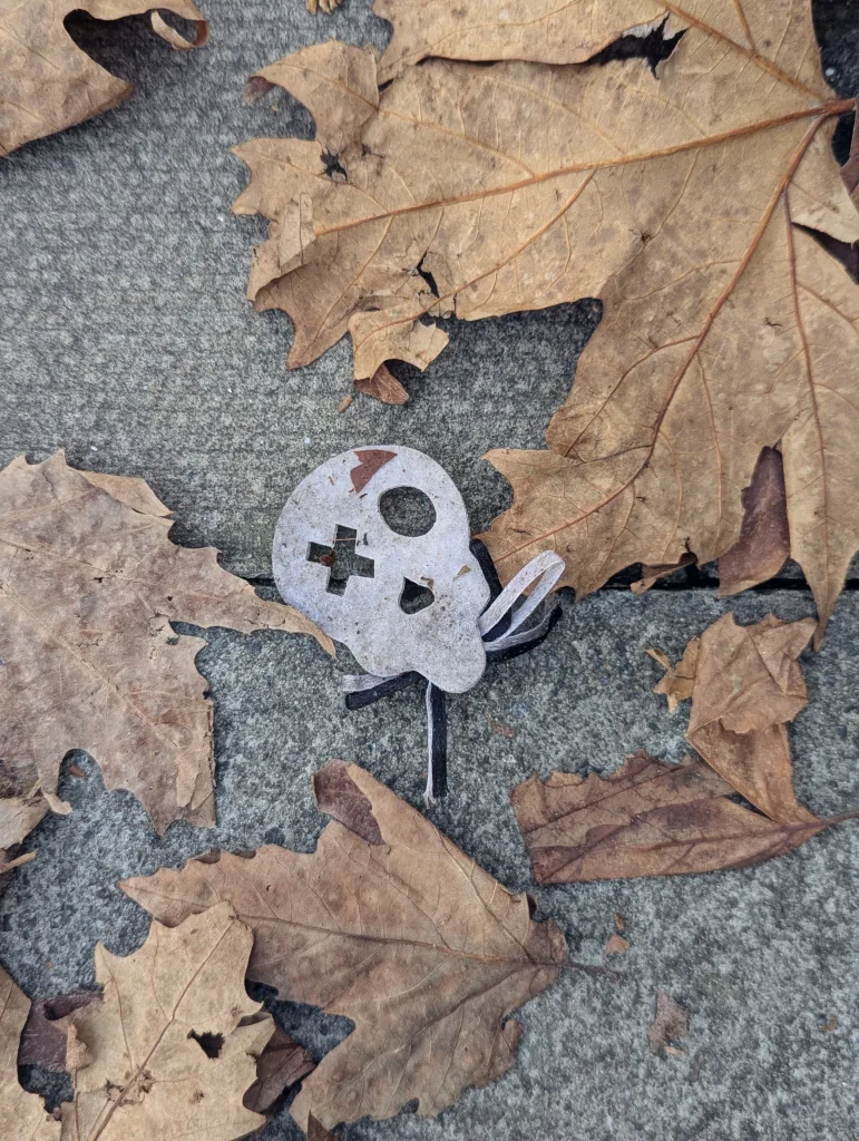 A white cardboard skull cutout with a plus-shaped eye lying among dry brown autumn leaves on a stone pavement.