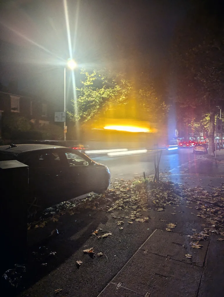 Long exposure of yellow and white light trails on a misty street at night with fallen leaves on the pavement and a bright, flaring street lamp.