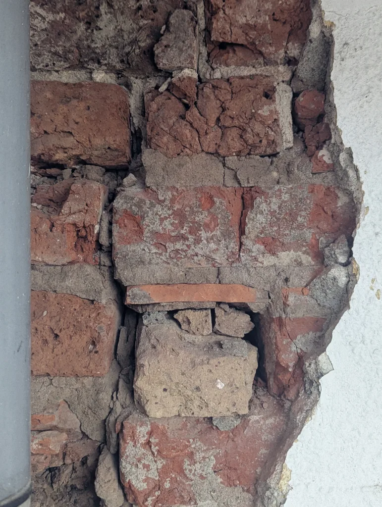 Exposed, crumbling red bricks and mortar behind a large crack in a white plastered wall beside a grey pipe.