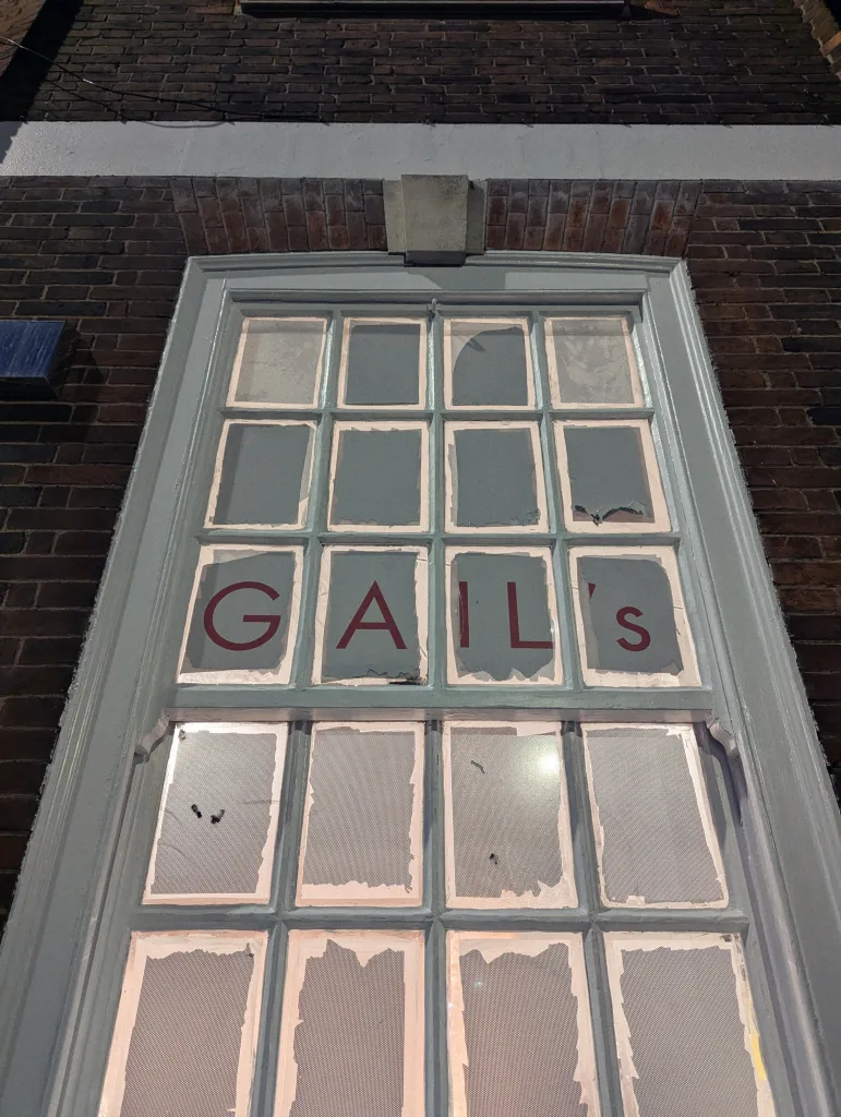Red GAIL’s lettering on a multi-pane window with peeling white film, set in a dark brick wall.