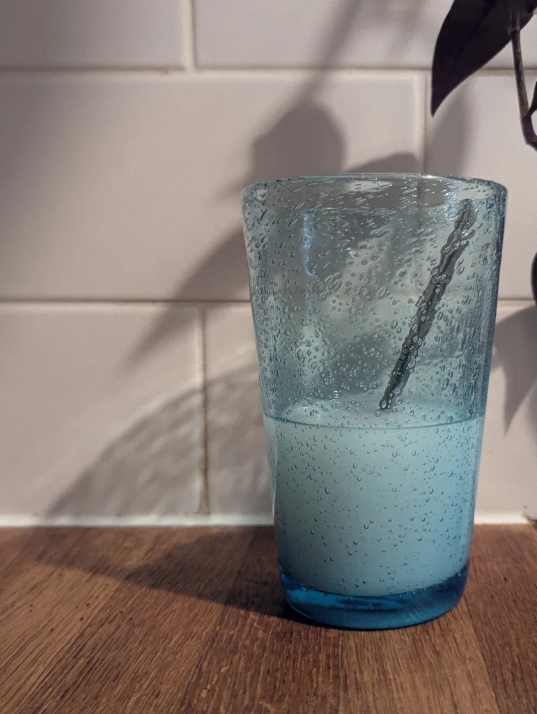 Opaque pale blue liquid in a bubbly blue glass with a metal stirrer, set on a wooden counter against white kitchen tiles.