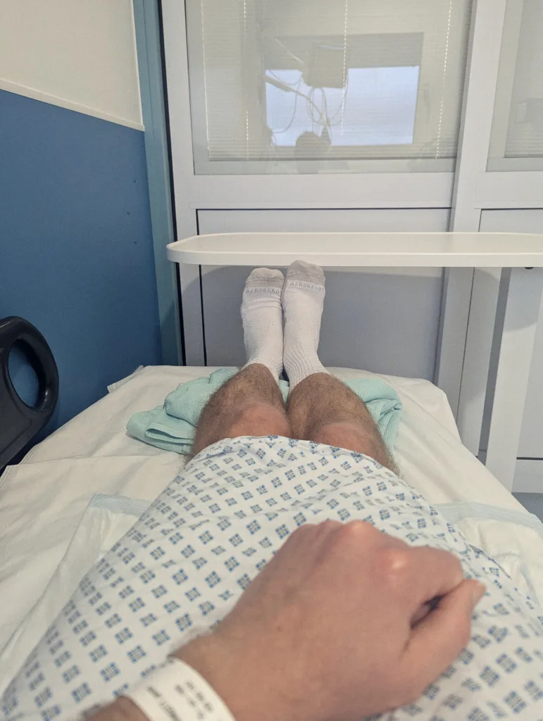 Patient's view from a hospital bed, showing legs in white Adidas socks, a blue patterned hospital gown, and a white ID wristband.