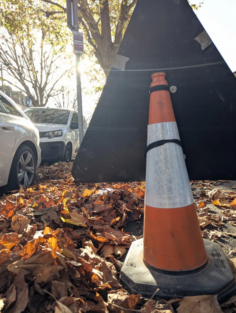 An orange traffic cone supporting a large black triangular sign on a pavement covered in autumn leaves in bright sunlight.