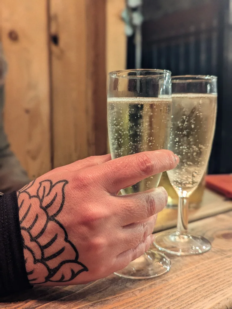 A hand with a floral tattoo holds a glass of bubbling wine next to another flute on a wooden table in a dimly lit setting.