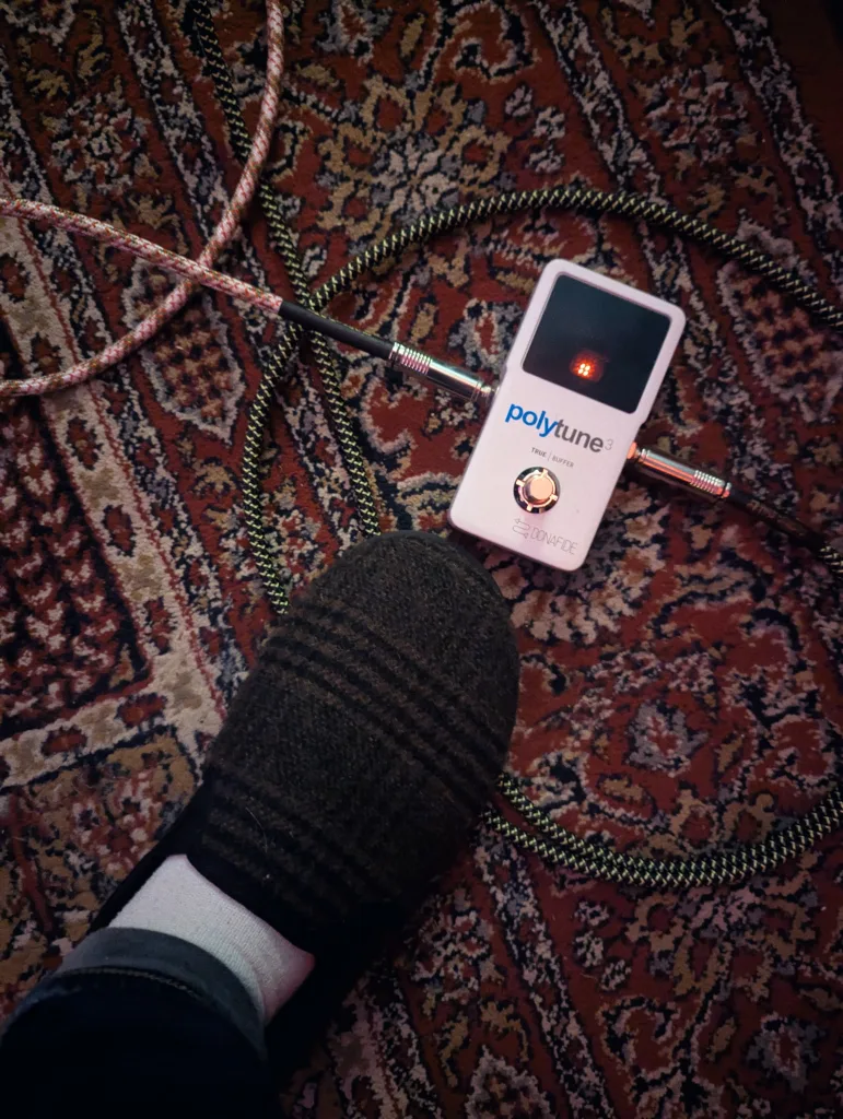 A white PolyTune 3 guitar pedal on an ornate red rug, with a foot in a patterned slipper and coiled braided cables.