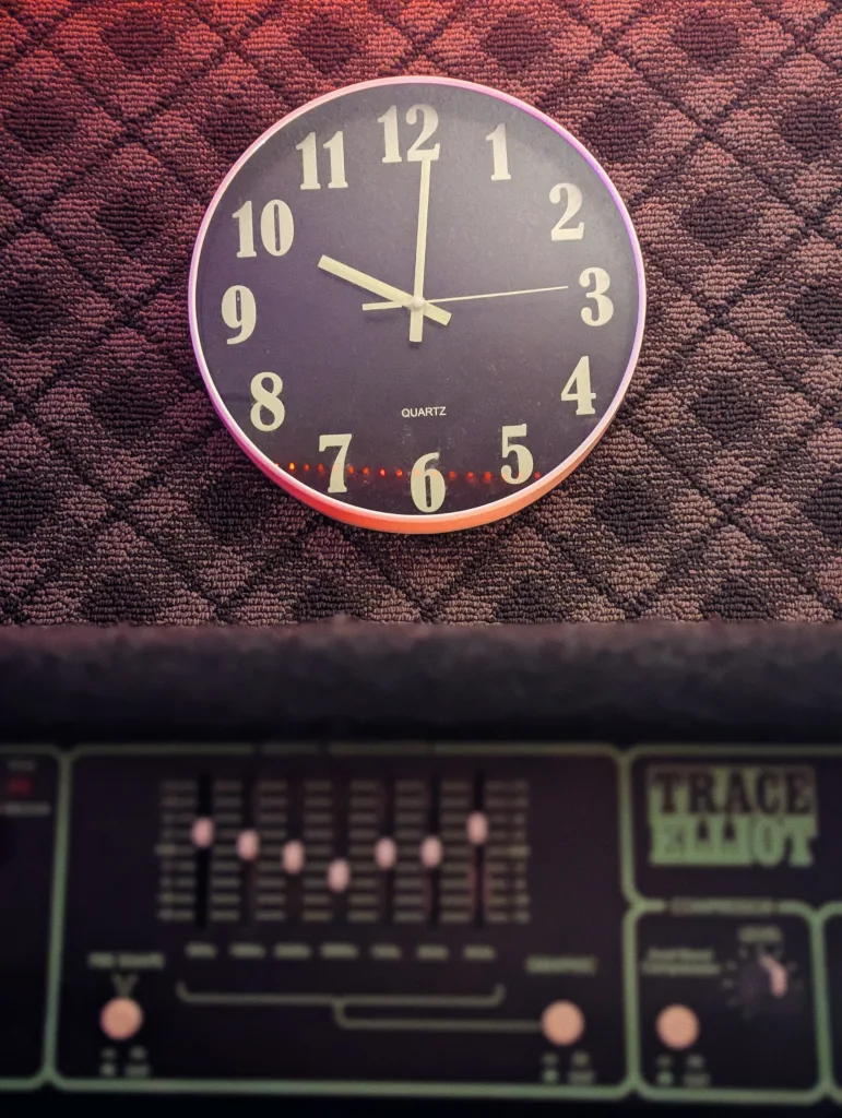 A quartz wall clock showing 10:02 on a diamond-patterned fabric wall above a Trace Elliot bass amplifier.