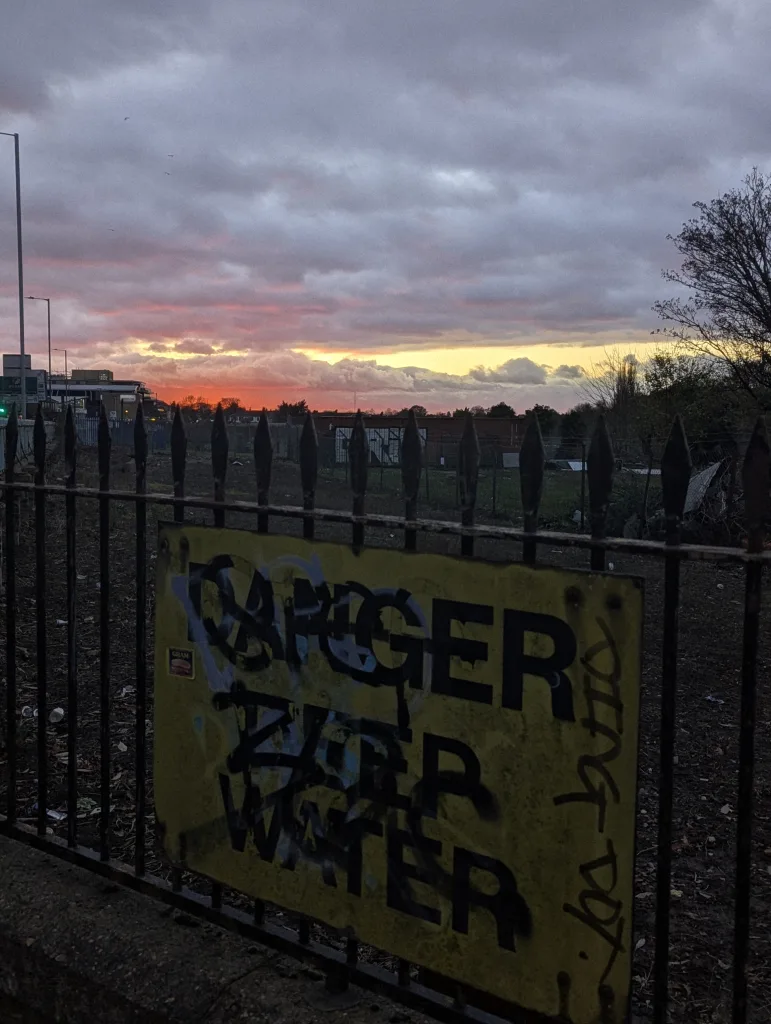 A graffitied yellow DANGER DEEP WATER sign on a spiked metal fence against a vibrant pink and orange sunset sky.