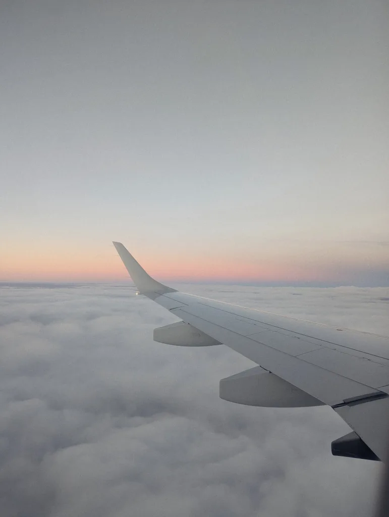 An airplane wing over a sea of white clouds with a thin band of pink sunset light on the horizon.