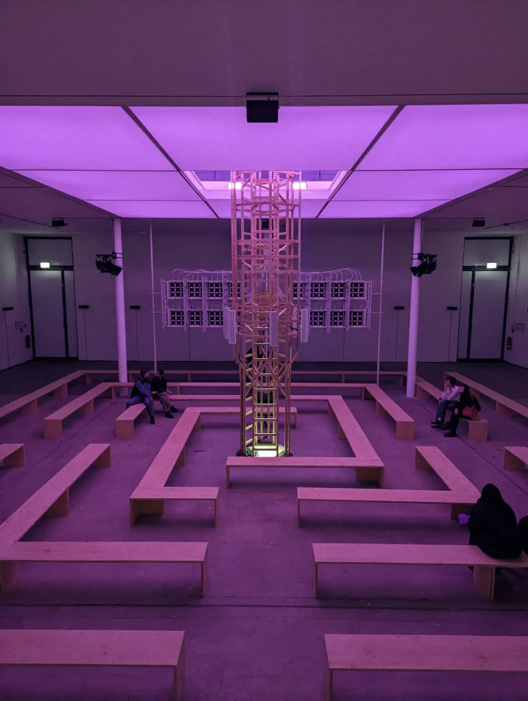 A room bathed in purple light with a central wooden tower installation and a labyrinth of benches where people sit.