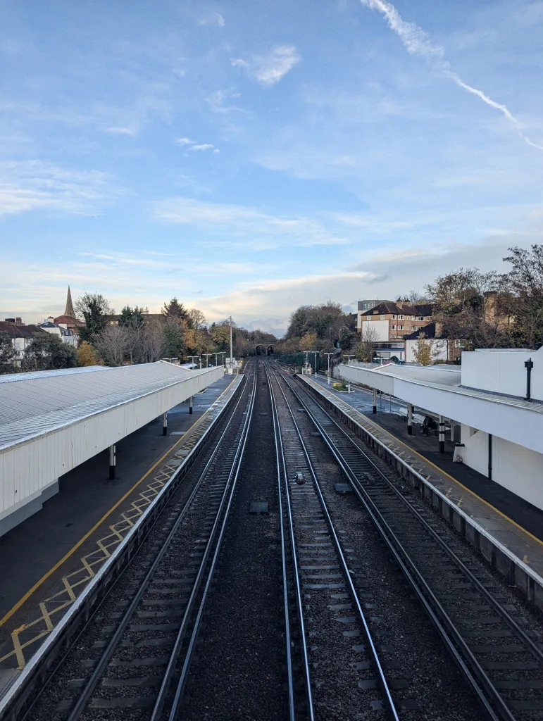 Straight railway tracks receding between two station platforms with white awnings under a blue sky with a long contrail.