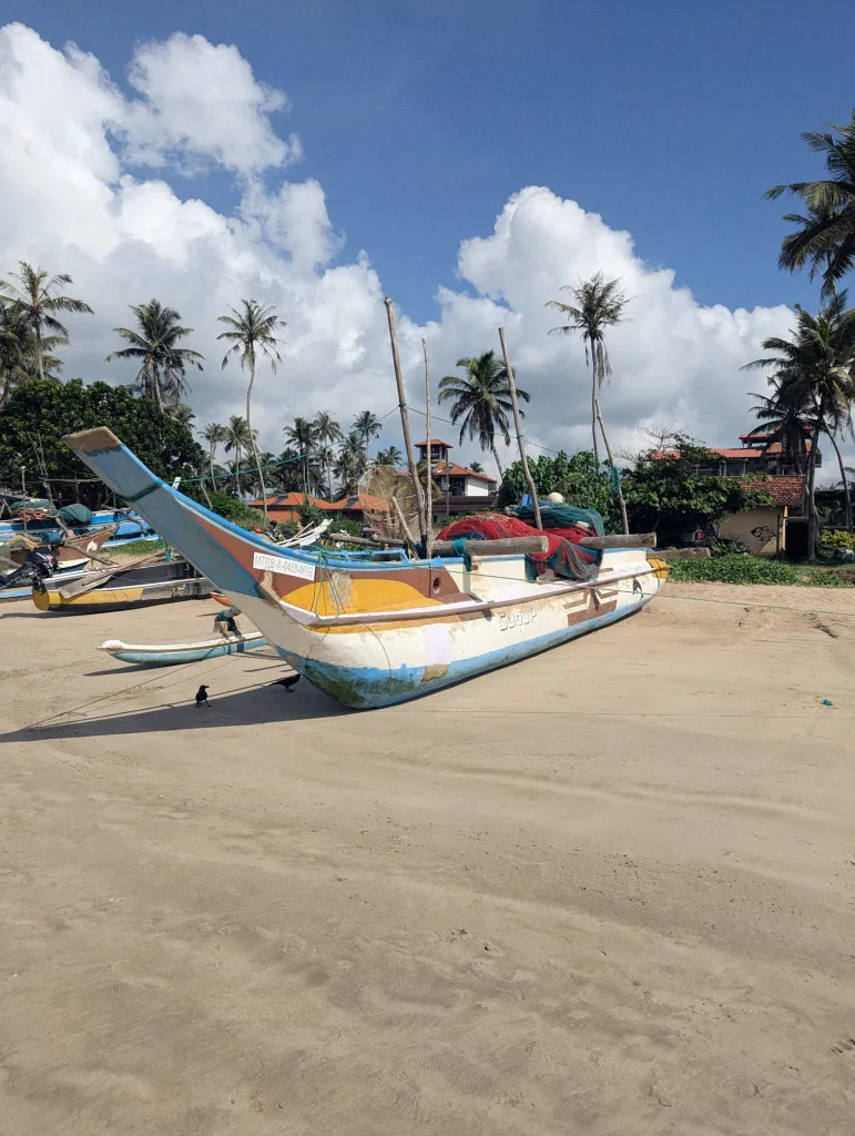 A vibrantly painted Sri Lankan outrigger fishing boat beached on sand, with palm trees and fluffy white clouds in the background.