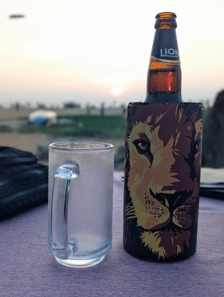 A Lion beer bottle in a lion-themed stubby holder next to a chilled glass mug on a table at sunset with a beach background.