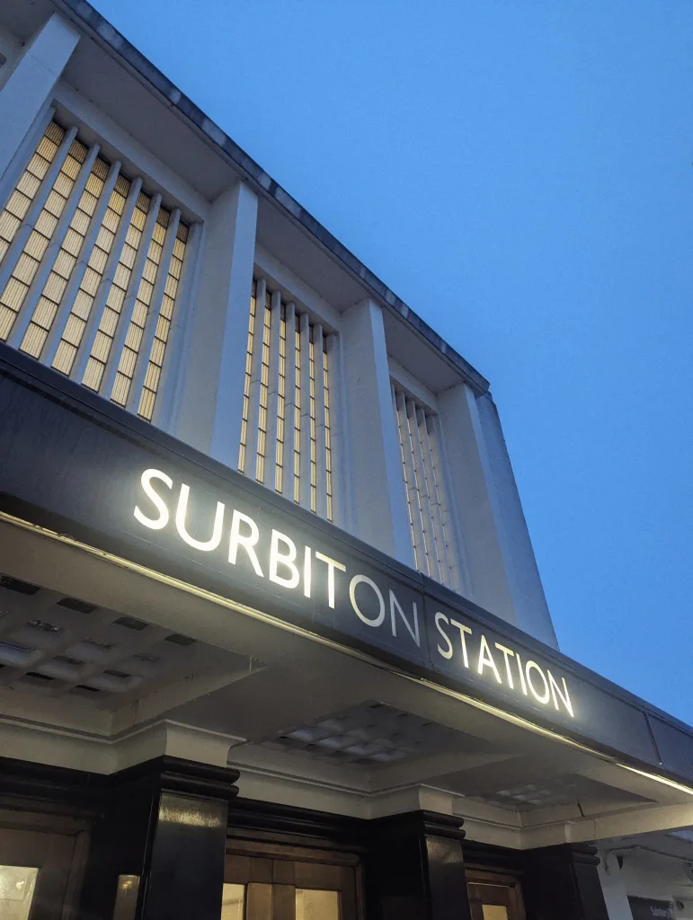 A glowing white sign for Surbiton Station on the art deco facade of the building against a deep blue dusk sky.