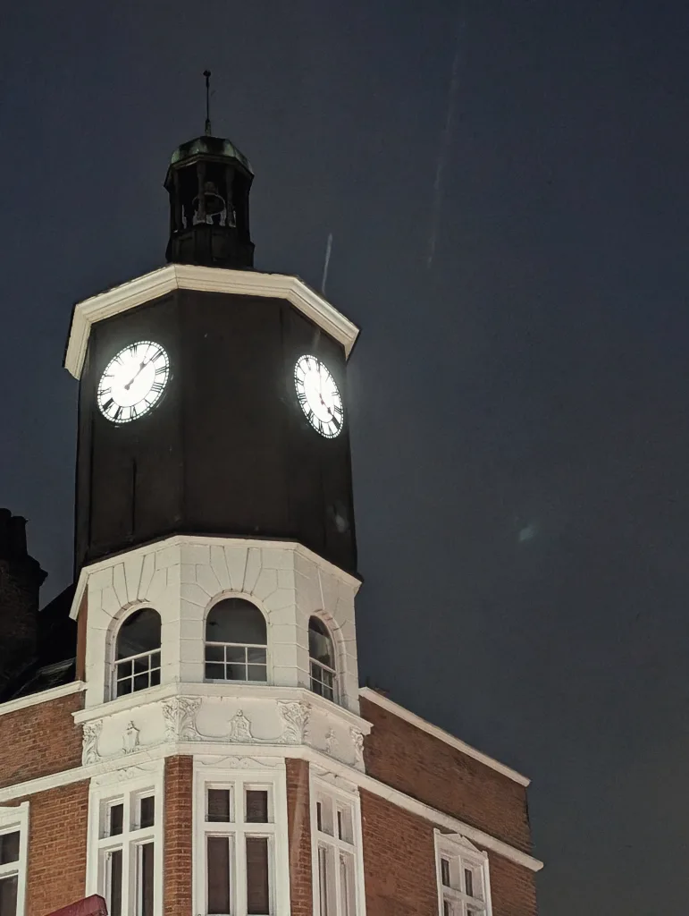 Two glowing white clock faces on a dark octagonal tower at night, atop a red brick building with ornate white stone details and arched windows.