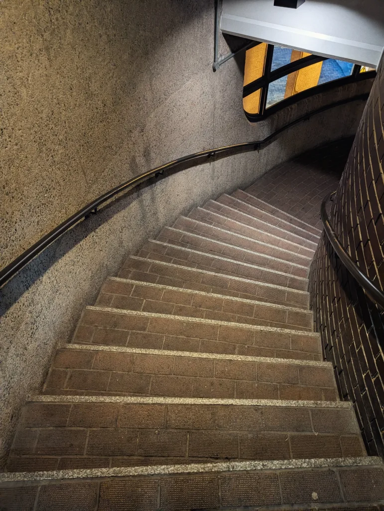 Brown brick steps curve downwards between a grey concrete wall and dark brickwork, framed by metal handrails in a dim architectural space.