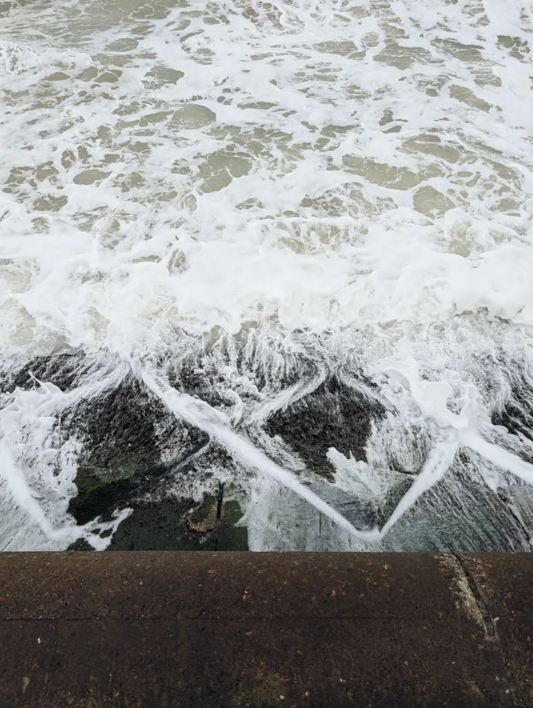 Frothy white waves and sea foam churning over dark, mossy rocks, viewed from above a brown stone sea wall.