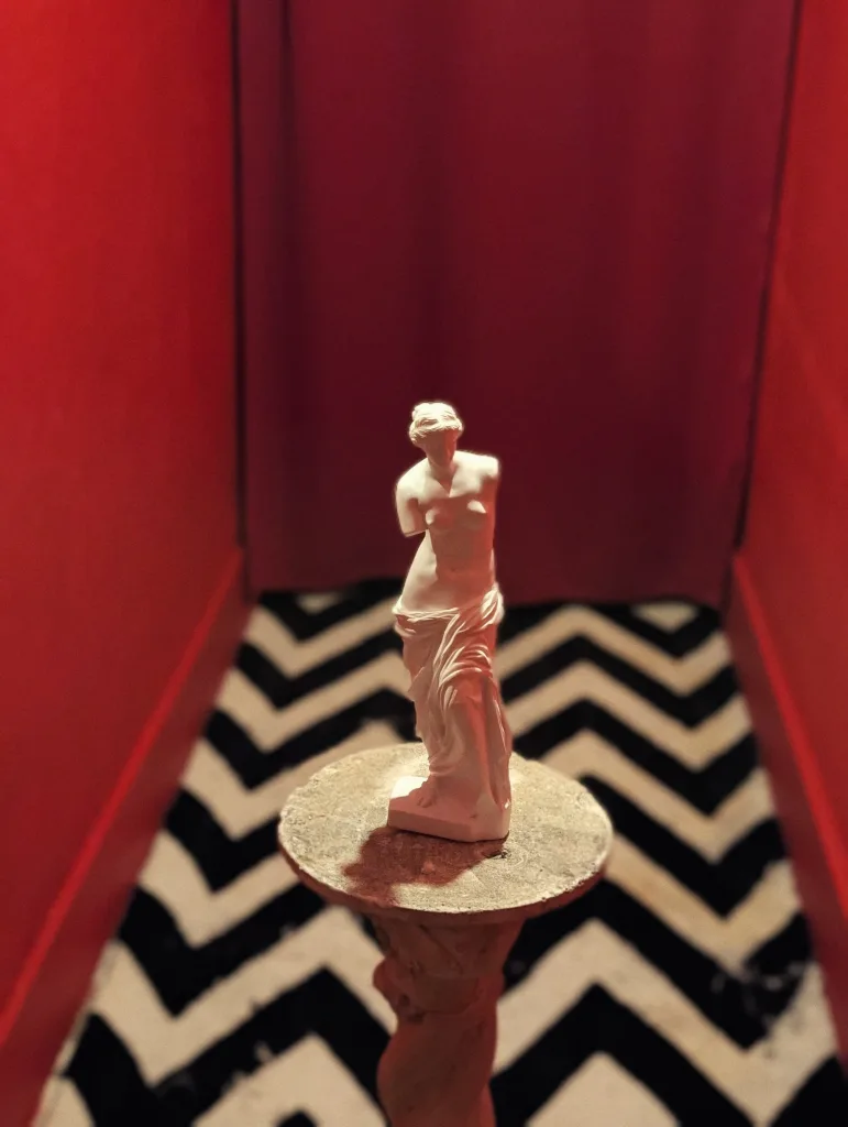 A miniature Venus de Milo statue on a pedestal against red curtains and a black-and-white chevron-patterned floor.