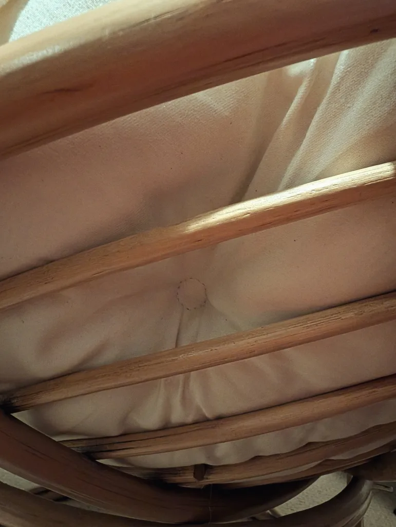 A cream-colored button-tufted cushion viewed through the parallel wooden poles of a curved papasan chair frame.