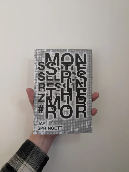 A hand holding the book MONSTERS IN THE MIRROR by Jay Springett, featuring bold black overlapping typography on a halftone cover against a white wall.
