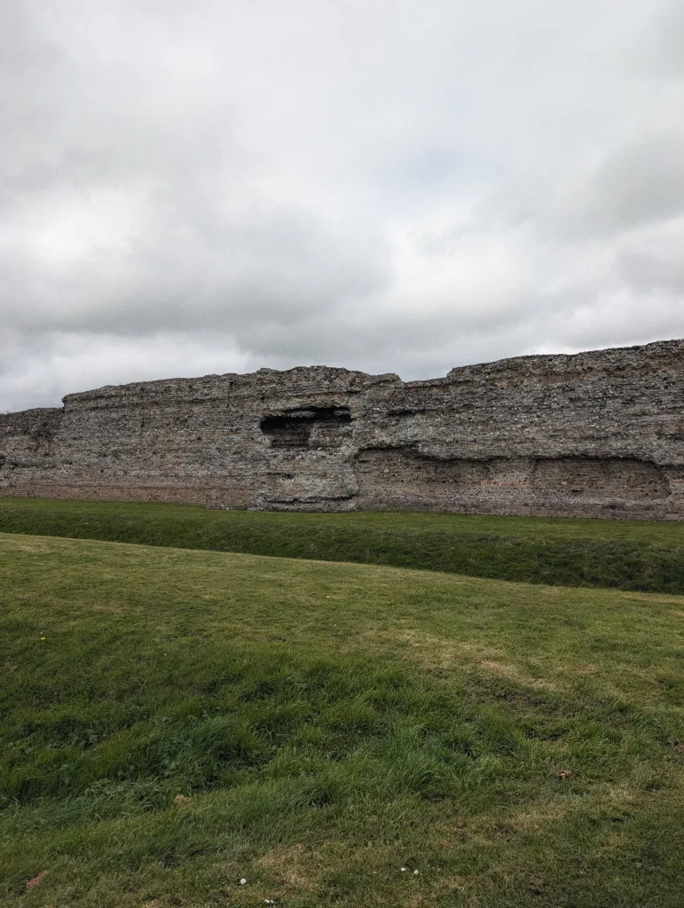 Ancient weathered Roman stone wall ruins stretch across a grassy field with earthwork ditches under an overcast grey sky.